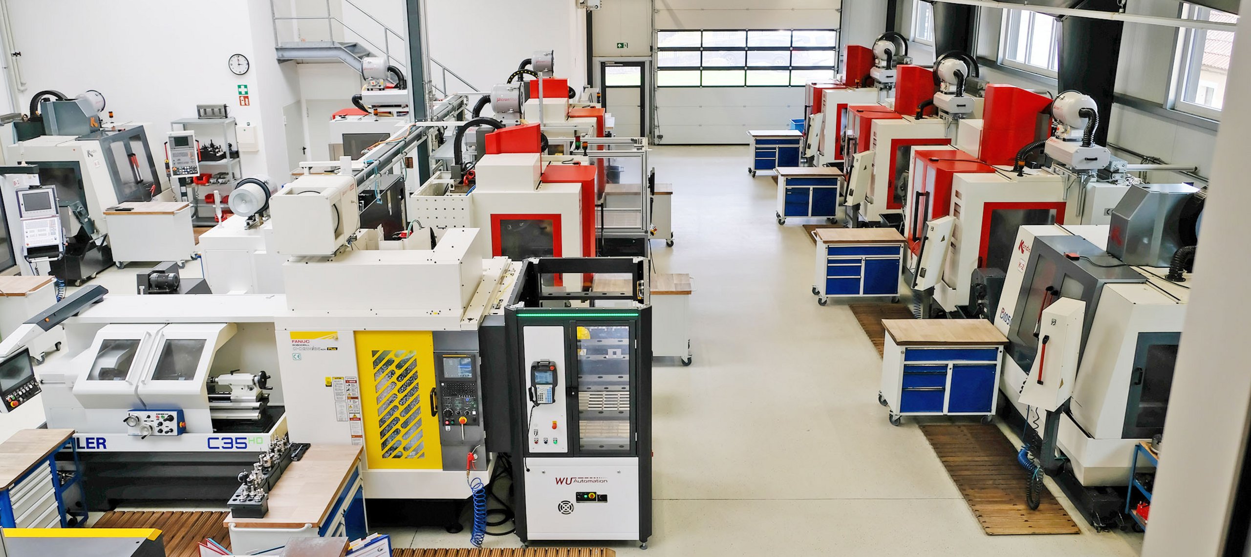 CNC- Zerspanung, WU-System Blick in eine Werkhalle mit mehreren CNC-Fräsmaschinen in Reihen, Arbeitsplätze mit blauen Schubladenschränken, helle Wände und große Fenster.